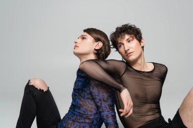 Gri üzerinde tek başına otururken eşine yaslanan siyah şeffaf üstlü moda biseksüel modeli