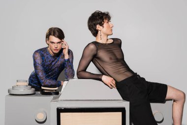 Siyah şeffaf bluzlu genç çift cinsiyetli kişi gri arka planda, modaya uygun ortağın yanında büyük bir fotoğraf makinesi modelinin üzerinde oturuyor.