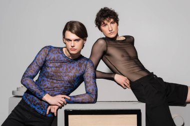 nonbinary couple in black and animal print tops leaning on huge model of photo camera on grey background clipart