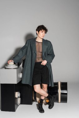 Gri arka planda fotoğraf makinesi modelinin yanından uzaklaşan ceketli ve siyah deri çizmeli şık olmayan tam boy.