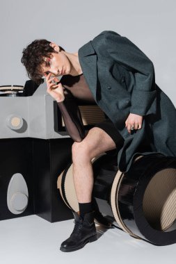 Düşünceli çift cinsiyetli insan gri arkaplanda fotoğraf makinesinin büyük bir modelinin üzerinde oturuyor.