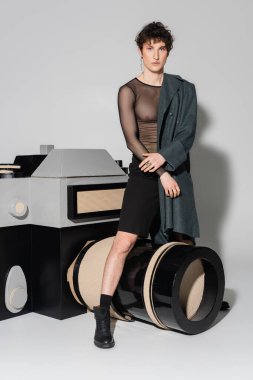 Gri arkaplanda fotoğraf makinesinin büyük bir modelinin yanında duran siyah şeffaf bluzlu ve ceketli modaya uygun olmayan kişi.