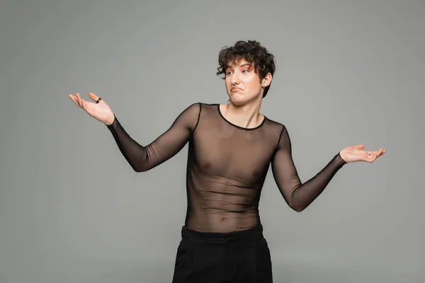 confused pangender person in black transparent top showing shrug gesture isolated on grey