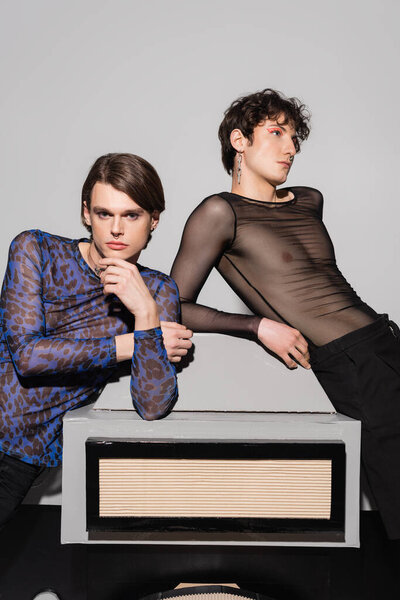 young and stylish pansexual partners posing near big model of photo camera on grey background