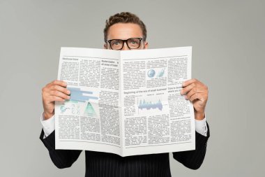 Gözlüklü bir işadamı kameraya bakarken yüzünü gazeteyle kapatıyor. 
