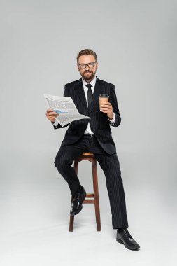 full length of happy businessman in suit and glasses holding newspaper while sitting with paper cup on grey  clipart