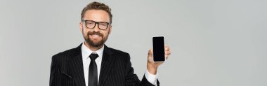 Takım elbiseli ve gözlüklü neşeli işadamı gri pankartta boş ekranlı akıllı telefonu gösteriyor. 