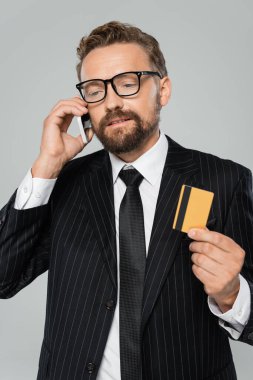 Takım elbiseli ve gözlüklü sakallı işadamı akıllı telefondan konuşuyor ve kredi kartını gri renkte tutuyor. 