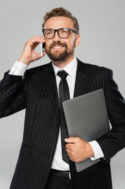 Gözlüklü pozitif işadamı dizüstü bilgisayarı tutuyor ve gri telefonda konuşuyor. 
