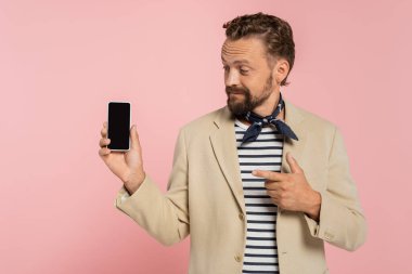 Spor ceket ve boyun atkısı takmış sakallı bir Fransız. Akıllı telefonu işaret ediyor. Pembe ekranda boş ekranı var. 