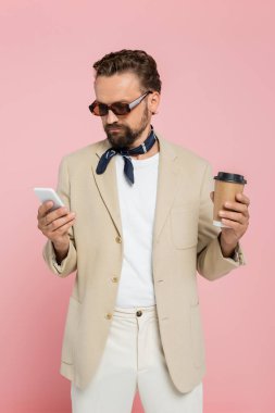 Boyunluk ve güneş gözlüklü şık bir Fransız adam kahve tutuyor ve pembe üzerine izole edilmiş akıllı telefon kullanıyor. 
