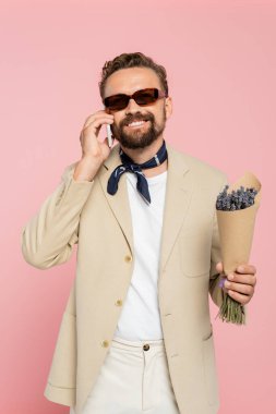 happy man in stylish sunglasses and neck scarf holding flowers and talking on smartphone isolated on pink   clipart