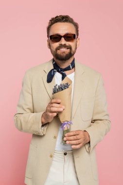 Şık güneş gözlüklü ve boynundaki atkıyla elinde çiçekleri pembe örtüyle gezen neşeli bir adam.  