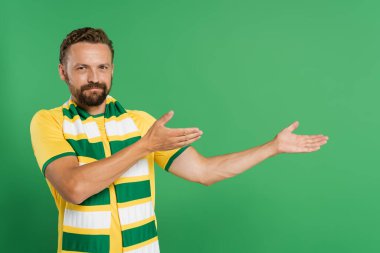 Çizgili eşarplı ve sarı tişörtlü sakallı futbol fanatiği yeşil ellerle kameraya bakarken kameraya bakıyor. 