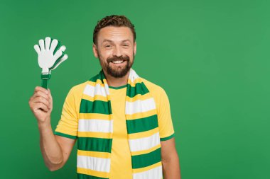 cheerful football fan in striped scarf holding plastic hand clapper isolated on green  clipart
