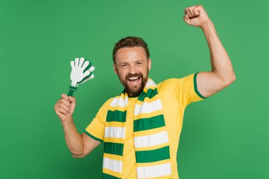 excited football fan in striped scarf holding plastic hand clapper isolated on green  clipart
