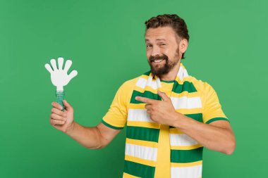 cheerful football fan in striped scarf pointing with finger at plastic hand clapper isolated on green  clipart