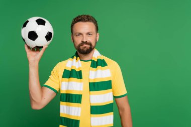 Çizgili eşarplı ve sarı tişörtlü futbol fanatiği yeşil üzerine izole edilmiş futbol topunu tutuyor. 
