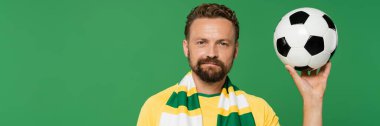 Çizgili eşarplı ve sarı tişörtlü sakallı spor fanatiği yeşil pankartta izole edilmiş futbol topu taşıyor. 