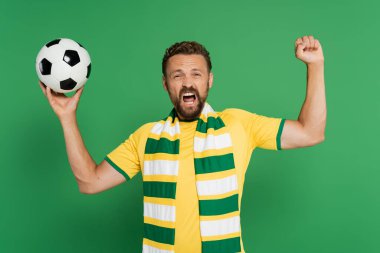 Çizgili eşarplı duygusal futbol fanatiği futbol topu tutuyor ve yeşilde izole edilmiş sevinçle eğleniyor 