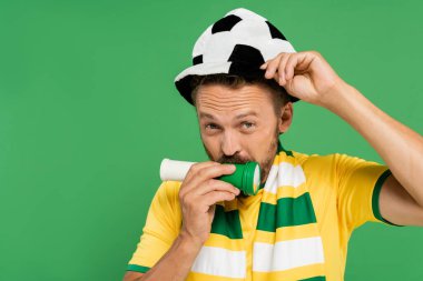 Futbol şapkalı sakallı adam yeşilde izole edilmiş kameraya bakarken korna çalıyor. 