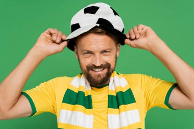Yeşil üzerine izole edilmiş futbol fanı şapkası takan çizgili atkı takan mutlu sakallı adam. 
