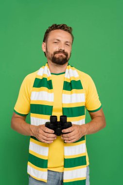 Futbol maçında elinde dürbün tutan çizgili fularlı sakallı bir adam yeşil alanda izole edilmiş.