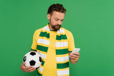 Çizgili eşarplı sakallı bir adam futbol topu tutuyor ve yeşil alanda izole edilmiş bir cep telefonu kullanıyor.