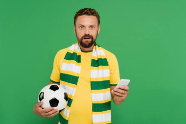 Yeşil üzerinde futbol topu ve cep telefonu tutan çizgili fularlı afallamış bir adam.