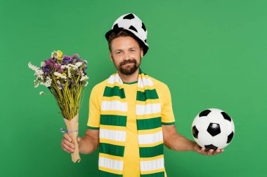 Spor şapkalı sakallı adam elinde futbol topu ve yeşil alanda yaban çiçekleri tutuyor. 