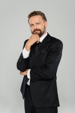 Resmi giyinmiş düşünceli ve sakallı bir adam gri kameraya bakıyor. 
