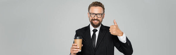 happy businessman in glasses and suit holding paper cup and showing thumb up isolated on grey, banner 