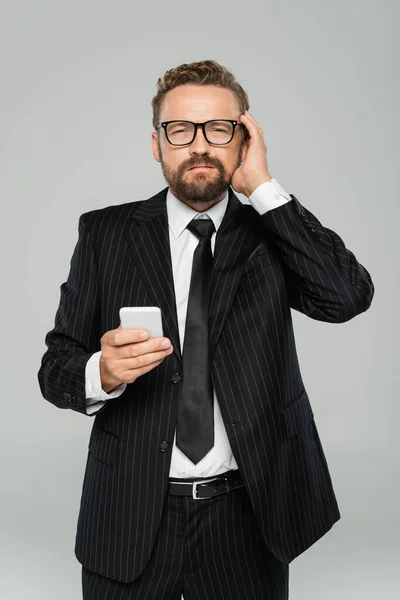 メガネやスーツの髭を生やしたビジネスマンが頭を悩ませながらグレーで隔離されたスマートフォンを持ち — ストック写真