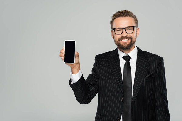 happy businessman in suit and glasses showing smartphone with blank screen isolated on grey 