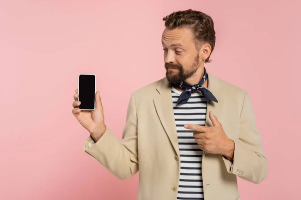 bearded french man in blazer and neck scarf pointing at smartphone with blank screen isolated on pink