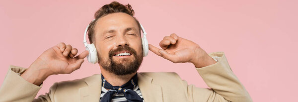 satisfied french man in blazer and neck scarf listening music in wireless headphones isolated on pink, banner 