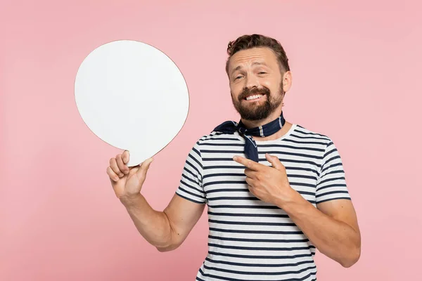 happy french man in striped t-shirt and neck scarf pointing at blank speech bubble isolated on pink