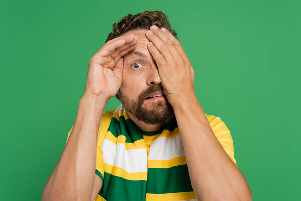 испуганный спортивный болельщик в полосатом шарфе и желтой футболке, покрывающий лицо изолированным зеленым 