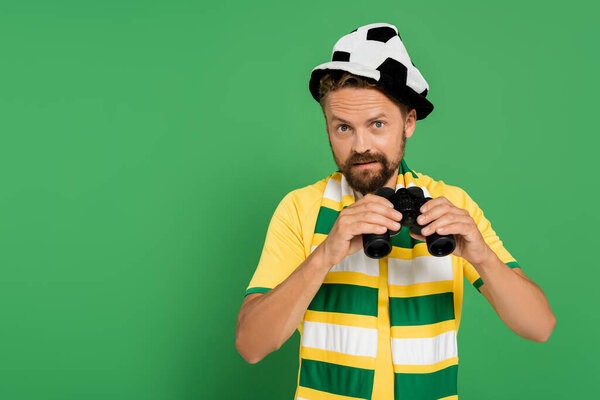 bearded man in fan hat and striped scarf holding binoculars during football match isolated on green 