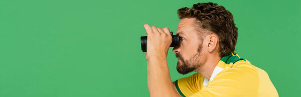 side view of bearded man in striped scarf holding binoculars during football match isolated on green, banner 