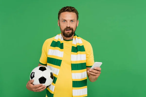 stunned man in striped scarf holding football and mobile phone isolated on green