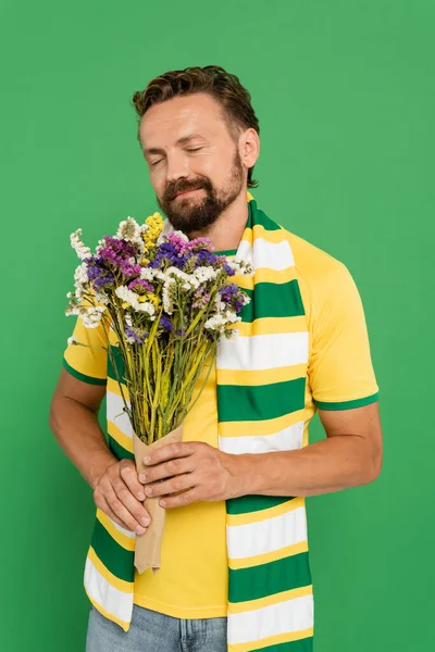 happy bearded football fan in scarf holding wildflowers isolated on green