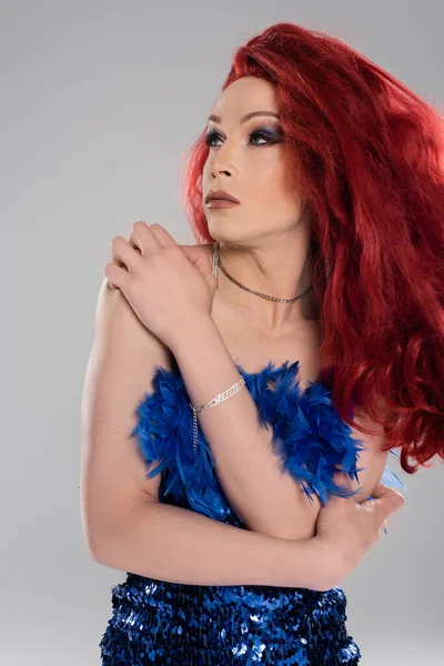 Transgender person in red wig and dress touching arm isolated on grey