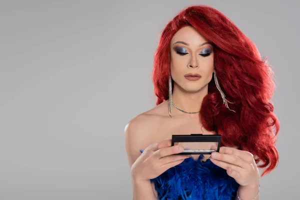 Elegant drag queen in red wig holding eye shadow isolated on grey