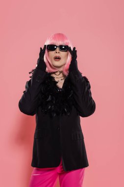 Peruklu ve ceketli bir travesti pembe arka planda güneş gözlüğüne dokunuyor. 