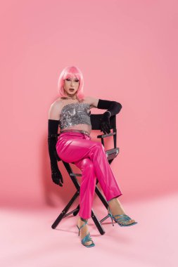 Parlak bluzlu moda travesti pembe arka planda sandalyede oturuyor. 