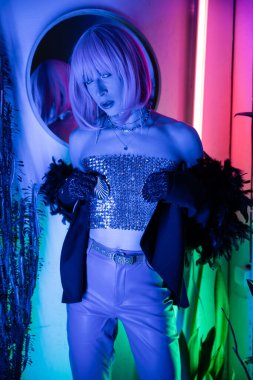 Moda travesti üstte ve ceketli. Evdeki neon ışıkta kameraya bakıyor. 