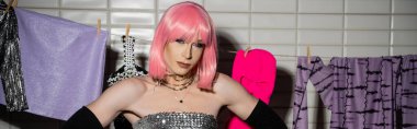 Pembe peruklu modaya uygun bir travesti banyoda iple bağlanmış kıyafetlerin yanındaki kameraya bakıyor. 