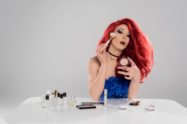 Fashionable drag queen applying face powder near decorative cosmetics and perfumes isolated on grey  