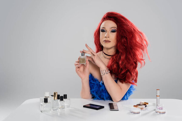 Drag queen in red wig holding perfume near decorative cosmetics isolated on grey  